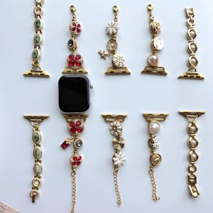 May include: An assortment of gold-toned Apple Watch bands with decorative charms. The bands feature various designs, including red and clear crystals, pearls, and floral accents. The watch bands are displayed on a light blue surface.