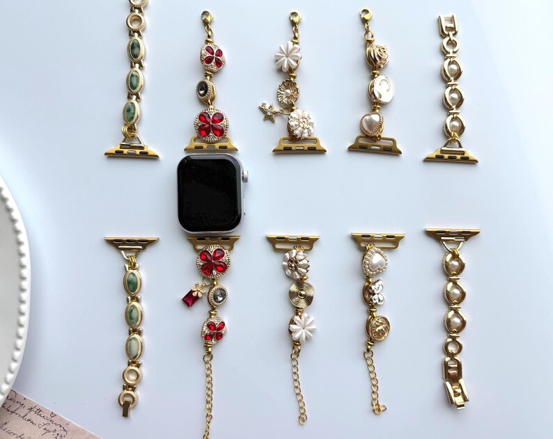 May include: An assortment of gold-toned Apple Watch bands with decorative charms. The bands feature various designs, including red and clear crystals, pearls, and floral accents. The watch bands are displayed on a light blue surface.