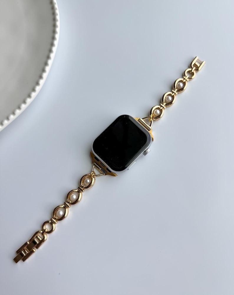 May include: A gold-toned watch band with a pearl and oval link design. The band is attached to a silver smartwatch with a black screen. The watch and band are on a white surface.