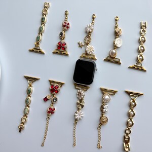 May include: An assortment of decorative watch bands in gold tones, adorned with gemstones, pearls, and floral accents. The bands feature red, green, and white embellishments, with a few including delicate chain details. A black smartwatch is also present.