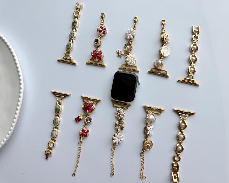 May include: An assortment of decorative watch bands in gold tones, adorned with gemstones, pearls, and floral accents. The bands feature red, green, and white embellishments, with a few including delicate chain details. A black smartwatch is also present.