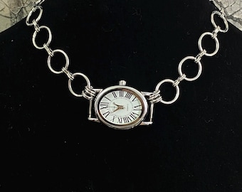 May include: A silver-toned necklace featuring a watch face as a pendant. The watch has a white face with black Roman numerals and hands. The chain is composed of large, open circular links.