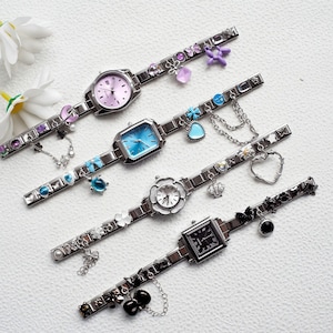 Op de afbeelding: Vier zilverkleurige polshorloges met decoratieve bedels. De horloges hebben verschillende gekleurde wijzerplaten, waaronder roze, blauw, wit en zwart. Bedels zijn onder meer bloemen, harten en andere decoratieve elementen. De horlogebanden zijn versierd met kleine, kleurrijke versieringen.