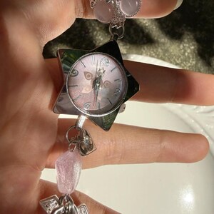 May include: A silver-toned watch with a star-shaped face and a pink dial. The watch is connected to a ring with pink stone accents and a dangling charm with pink beads. The watch has a delicate, feminine design.