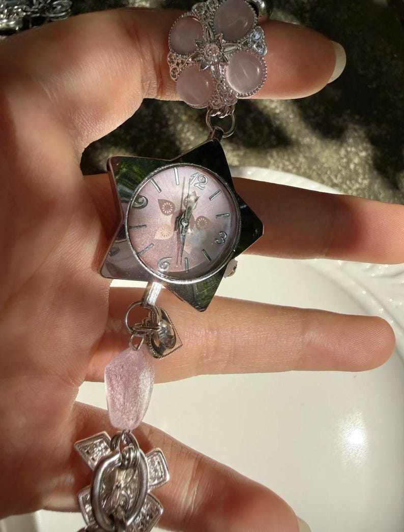 May include: A silver-toned watch with a star-shaped face and a pink dial. The watch is connected to a ring with pink stone accents and a dangling charm with pink beads. The watch has a delicate, feminine design.