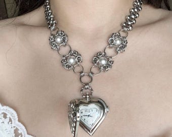 May include: Silver-toned necklace featuring a heart-shaped locket watch pendant. The necklace has a chain of small, round links and decorative floral elements with pearls. The watch face is white with black numerals and hands.