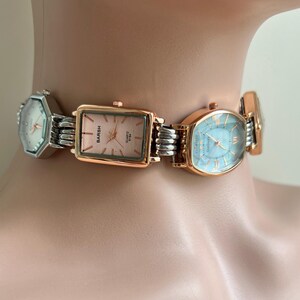 May include: A watch choker with multiple watch faces in silver and rose gold. The watch faces are various shapes, including rectangular and oval, with different colored faces. The choker is designed to be worn around the neck.