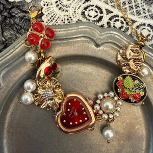 Peut inclure: Un bracelet doré orné d'une montre en forme de cœur avec un cadran rouge et des cristaux. Le bracelet est orné de breloques, notamment des perles rouges en forme de fleur, de fausses perles et un motif de fraise. Le cadran de la montre indique "Kapehuo".