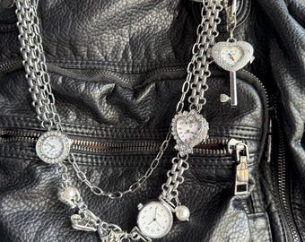 May include: A silver-toned necklace with multiple watch faces and charms, including a heart-shaped watch and a key pendant. The necklace is layered and adorned with faux pearls and is set against a black leather bag.