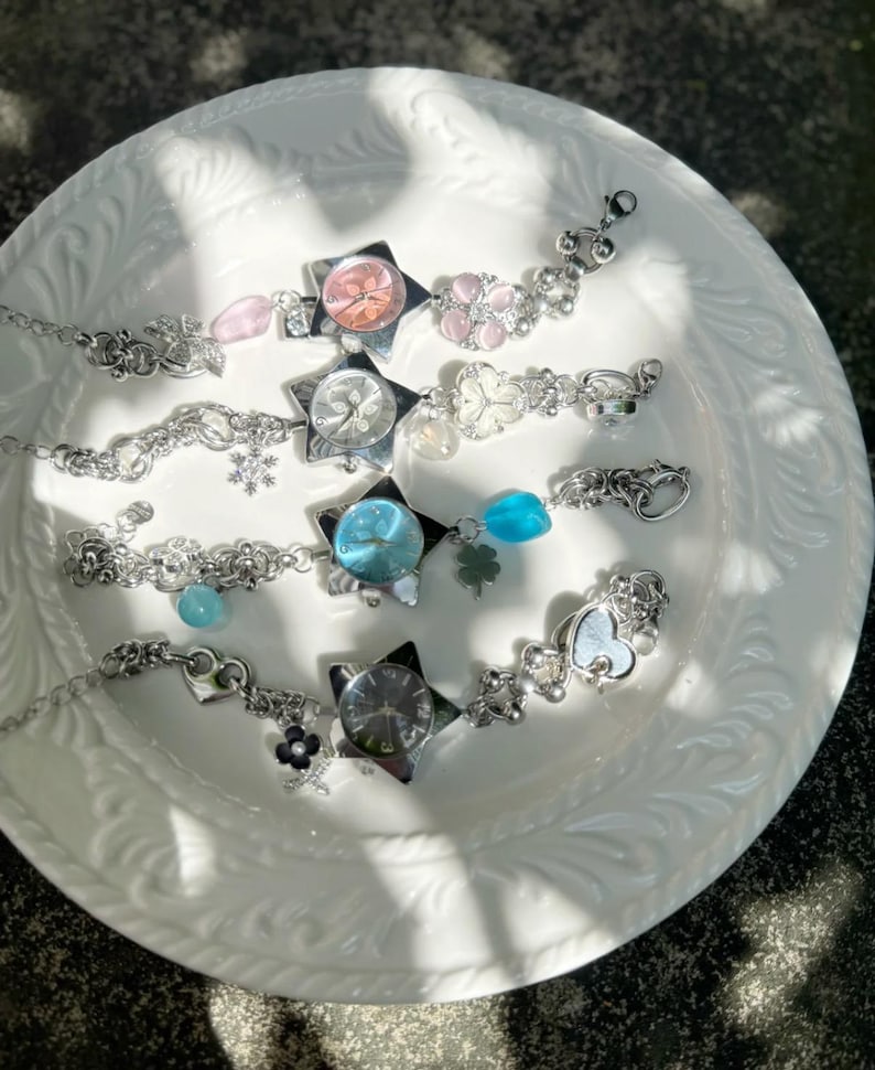 May include: Several silver-toned charm bracelets with various embellishments, including star-shaped watch faces, floral accents, and heart-shaped charms. The bracelets are displayed on a white, textured plate.