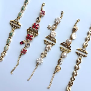 以下が含まれることがあります： さまざまな装飾要素を備えた5つのゴールドトーンのブレスレット。1つのブレスレットは緑色の楕円形の石を特徴とし、他のブレスレットは赤い花のアクセント、白い花、真珠、その他の装飾を特徴としています。各ブレスレットはユニークなデザインとチェーンクロージャーを備えています。