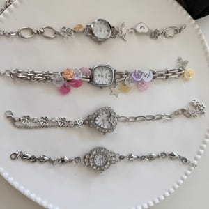 Może przedstawiać: Cztery srebrne zegarki na rękę z ozdobnymi paskami. Jeden ma okrągłą tarczę z kwiatowym i gwiazdkowym wzorem. Inny ma kwadratową tarczę z kolorowymi guzikami. Trzeci ma tarczę w kształcie serca, a czwarty okrągłą z kryształowym wzorem.