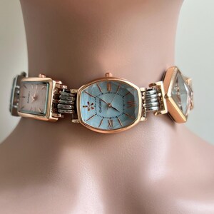 May include: A rose gold and silver watch choker with three watch faces. The central watch face is oval with a light blue face and Roman numerals. The other two watch faces are square and diamond-shaped. The watch is connected by silver links.