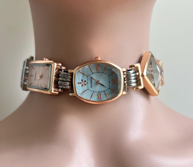 May include: A rose gold and silver watch choker with three watch faces. The central watch face is oval with a light blue face and Roman numerals. The other two watch faces are square and diamond-shaped. The watch is connected by silver links.