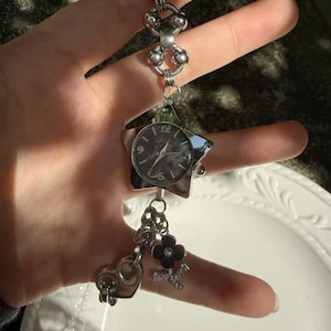 May include: A silver-toned watch bracelet with a star-shaped watch face and various charms. The charms include a black heart, a flower, and a letter "K". The watch face has a dark background with silver hands and numerals.