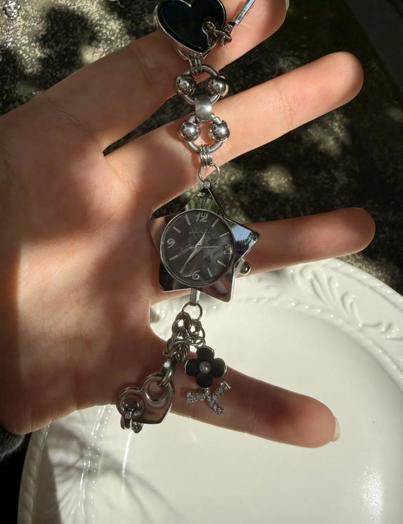 May include: A silver-toned watch bracelet with a star-shaped watch face and various charms. The charms include a black heart, a flower, and a letter "K". The watch face has a dark background with silver hands and numerals.