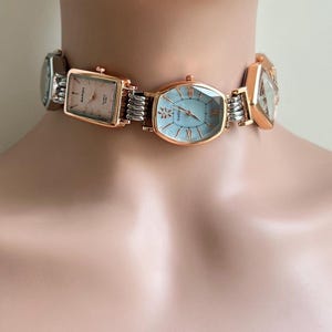 May include: A distinctive choker necklace composed of several watch faces in diverse shapes. The watches display silver and rose gold hues with light-coloured faces. Silver-toned metal links connect the watches, forming a bold accessory.