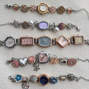 May include: A selection of decorative women's watches with diverse face shapes and adornments. The watches have silver-tone metal straps and colourful faces, including pink, blue, and white. Some watches feature floral and charm accents.