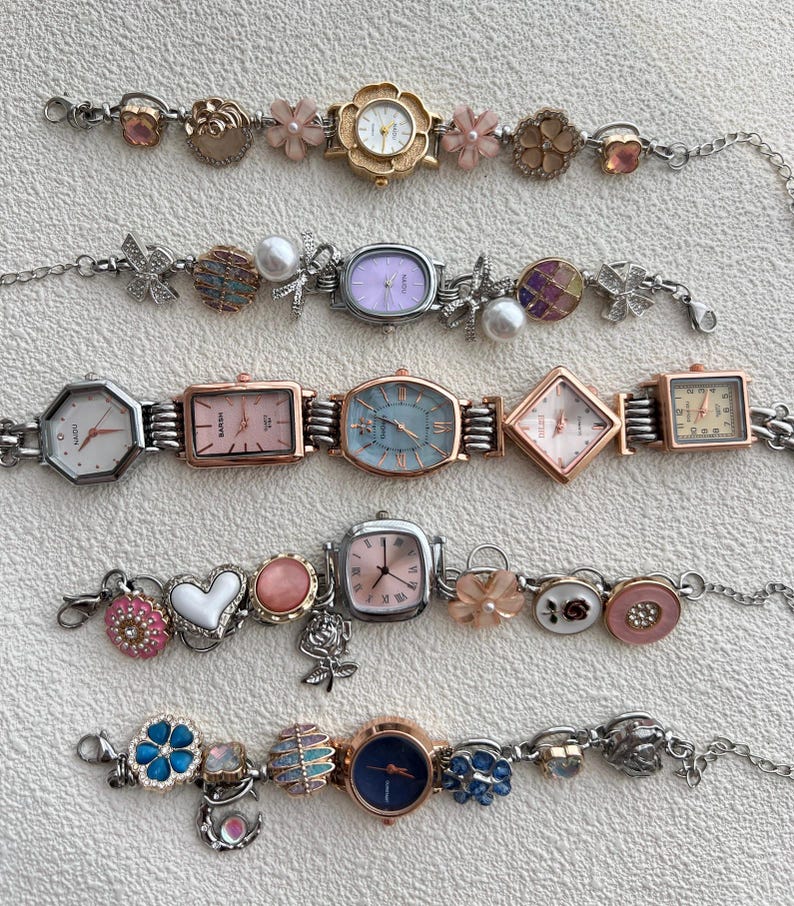 May include: A selection of decorative women's watches with diverse face shapes and adornments. The watches have silver-tone metal straps and colourful faces, including pink, blue, and white. Some watches feature floral and charm accents.