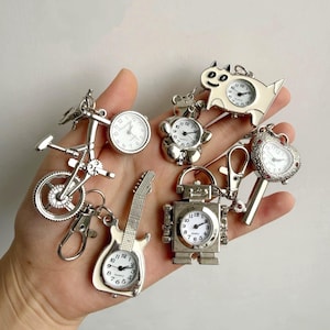 Puede incluir: Una colección de relojes en miniatura plateados de varias formas, incluyendo una bicicleta, una guitarra, un robot y un perro. Cada reloj tiene una esfera blanca con números y manecillas negras. Los relojes están unidos a llaveros.