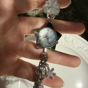 May include: Silver-toned watch with a star-shaped face and a white dial. The watch features a decorative chain with a pearl and a snowflake charm. The band includes a flower-shaped design. The watch is approximately 7 inches long.