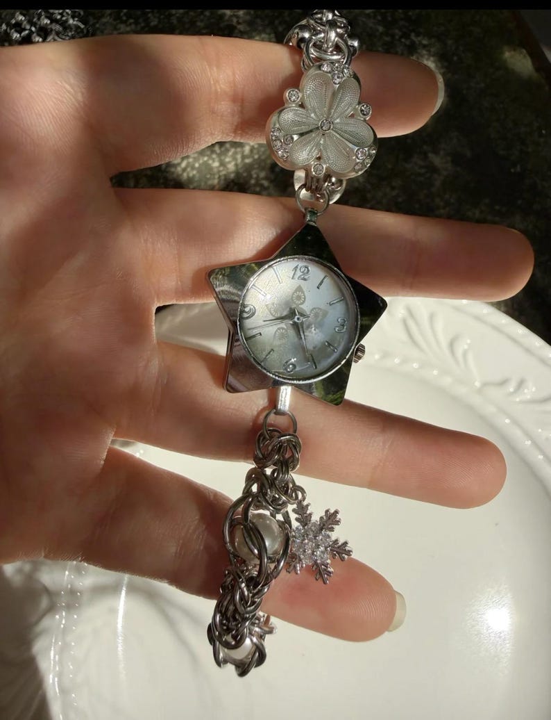 May include: Silver-toned watch with a star-shaped face and a white dial. The watch features a decorative chain with a pearl and a snowflake charm. The band includes a flower-shaped design. The watch is approximately 7 inches long.