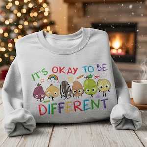 Peut inclure: Un sweat-shirt gris clair avec l'inscription brodée "IT'S OKAY TO BE DIFFERENT" en lettres colorées. Le motif comprend un arc-en-ciel et plusieurs personnages de dessins animés. Le sweat-shirt a des manches longues et un col rond.