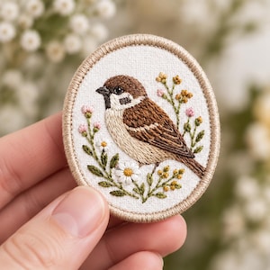 May include: An oval-shaped embroidered patch featuring a brown and white sparrow perched among small yellow and pink wildflowers on a white fabric background, finished with a tan stitched border around the entire edge of the piece.