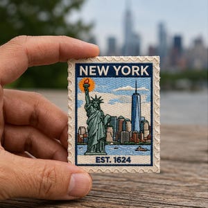 May include: A rectangular embroidered patch shaped like a postage stamp with a scalloped border. It features the Statue of Liberty, the New York City skyline, and the text NEW YORK at the top and EST. 1624 at the bottom in dark blue lettering.