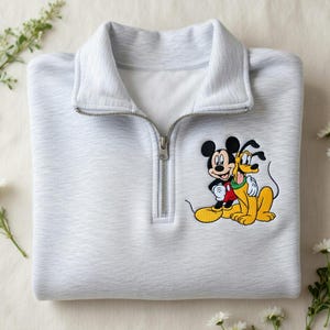 May include: A pale grey sweatshirt with a quarter-zip collar. The sweatshirt has an embroidered image of Mickey Mouse and Pluto. The characters are in their classic colours: black, red, yellow, and white. The sweatshirt is neatly folded.