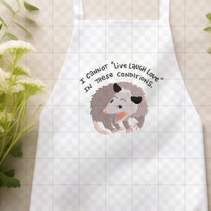 Peut inclure: Tablier blanc avec un dessin d'opossum de dessin anim&eacute; et le texte "I cannot 'Live Laugh Love' in these conditions." L'opossum est gris, rose et noir. Le tablier a un bavoir et des liens &agrave; la taille.