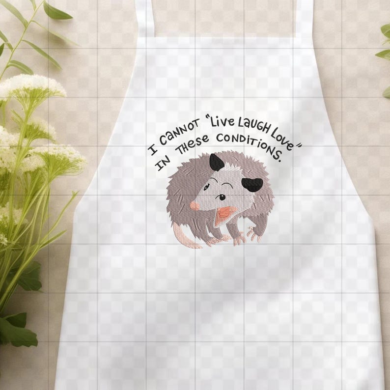 Peut inclure: Tablier blanc avec un dessin d'opossum de dessin anim&eacute; et le texte "I cannot 'Live Laugh Love' in these conditions." L'opossum est gris, rose et noir. Le tablier a un bavoir et des liens &agrave; la taille.