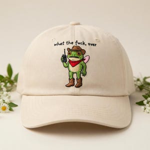 May include: Cream-colored baseball cap with the embroidered phrase "what the fuck, over." The cap features an embroidered cartoon frog wearing a cowboy hat, red bandana, and boots, holding a walkie-talkie.