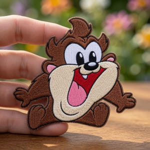 May include: An embroidered iron-on patch featuring the cartoon character Taz, the Tasmanian Devil. The design shows the brown-furred character with a wide, open mouth, a pink tongue, white eyes, and a black nose, rendered in detailed thread work.