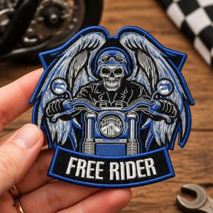 May include: An embroidered patch featuring a skull wearing a helmet and goggles, riding a motorcycle with large wings extending from the sides. The design uses blue, black, and white thread, with the text FREE RIDER printed on a banner at the bottom.
