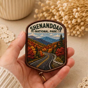 May include: A rectangular embroidered patch featuring a scenic landscape of Shenandoah National Park. The design depicts a winding road through a forest with autumn-colored trees, blue mountains in the distance, and the text Shenandoah National Park.