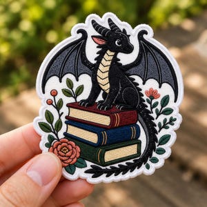 May include: An embroidered patch featuring a black dragon with pink-lined ears perched atop a stack of three books in red, blue, and green. The design is framed by green leaves, small orange berries, and a large peach-colored flower on a white background.
