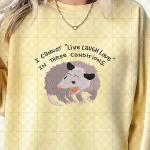 Peut inclure: Sweat-shirt jaune clair avec l'inscription "I CANNOT 'Live Laugh Love' IN THESE CONDITIONS." Un opossum de dessin anim&eacute; avec une fourrure grise, une bouche rose et des oreilles noires est centr&eacute; sous le texte. Le sweat-shirt a un col rond et des manches longues.