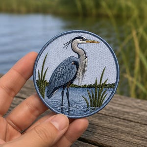 May include: A circular embroidered patch featuring a detailed depiction of a great blue heron standing in water among green reeds. The bird has grey and blue plumage, a long neck, and a yellow beak. The patch has a blue stitched border.