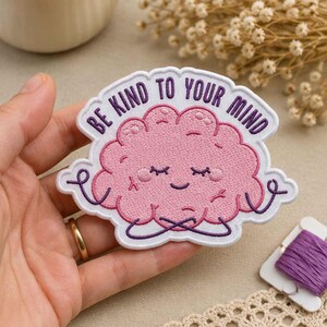 Può includere: Una toppa ricamata raffigurante un personaggio a forma di cervello rosa con un viso sorridente e gli occhi chiusi, seduto a gambe incrociate. Il testo BE KIND TO YOUR MIND è ricamato in viola sopra il cervello su sfondo bianco con bordo smerlato.
