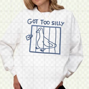 May include: White sweatshirt with a blue graphic of a bird in a cage, with the text "GOT TOO SILLY" above the cage. The design is a simple line drawing style.