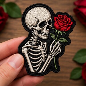 Peut inclure: Un écusson brodé représentant un torse et un crâne de squelette beige tenant une rose rouge éclatante avec des feuilles vertes. Le motif est souligné par un fil noir, mettant en valeur les détails de la cage thoracique et du visage.