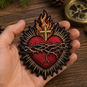 May include: An embroidered patch featuring a red heart encircled by a brown crown of thorns, topped with a gold cross and orange flames, all surrounded by radiating gold rays with a black border, presented as a detailed textile emblem.