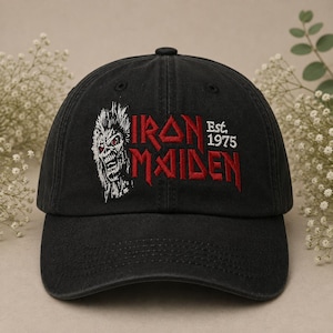 Puede incluir: Una gorra de béisbol negra con un bordado de la mascota de Iron Maiden, Eddie, en blanco y rojo, junto con el texto rojo Iron Maiden y el texto blanco Est. 1975 bordados en el panel frontal de la corona estructurada de seis paneles.
