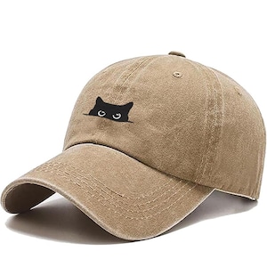 May include: A tan baseball cap with a curved brim. The cap features a black cat graphic peeking over the top. The hat is made of a soft, textured material and has a classic six-panel design.