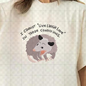 Peut inclure: T-shirt cr&egrave;me avec un opossum de dessin anim&eacute; et le texte "I CANNOT 'Live Laugh Love' IN THESE CONDITIONS." L'opossum est gris avec des oreilles noires et une bouche rose. Le t-shirt a une coupe ample.