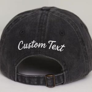May include: A black baseball cap with the text "Custom Text" embroidered in white. The cap has a curved brim and an adjustable strap with a metal buckle.
