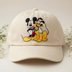 Puede incluir: Gorra de béisbol beige con una imagen bordada de Mickey Mouse y Pluto. Mickey lleva una camisa roja y pantalones cortos negros, Pluto es amarillo. La gorra tiene una visera curva y un botón en la parte superior.