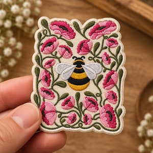 May include: A rectangular embroidered patch featuring a central bumblebee with yellow and black stripes and light gray wings, surrounded by a border of dark green vines and vibrant pink flowers on an off-white fabric background.