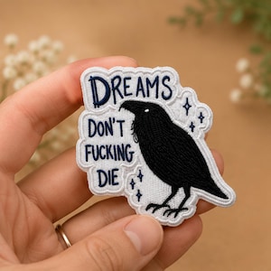 Könnte beinhalten: Ein bestickter Aufnäher mit weißem Hintergrund und gewelltem Rand. Er zeigt eine schwarze Rabenillustration und den dunkelblauen Text "DREAMS DON'T FUCKING DIE" in gestapelter Anordnung. Kleine dunkelblaue Sternsymbole sind um den Raben verteilt.